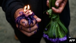 Un devoto sostiene una vela encendida en su mano y un ramo de flores moradas, en tributo a San Lázaro.