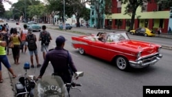 Turistas pasean en un almendrón mientras varias personas esperan por el transporte público en una parada en La Habana.
